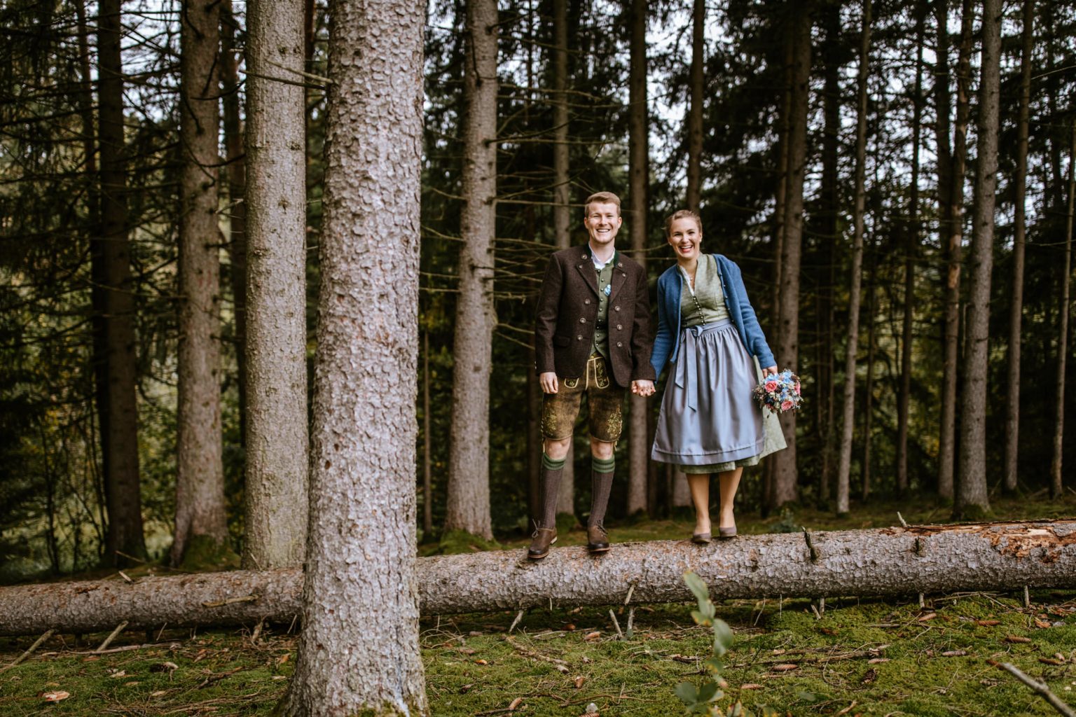 Hochzeitspaar in Tracht steht auf einem Baumstamm im Wald