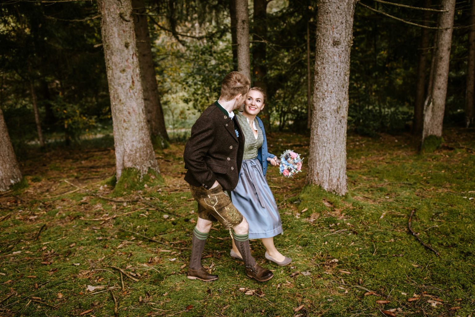 Bräutigam läuft mit seiner Braut in Tracht durch den Wald in der Nähe von Weilheim